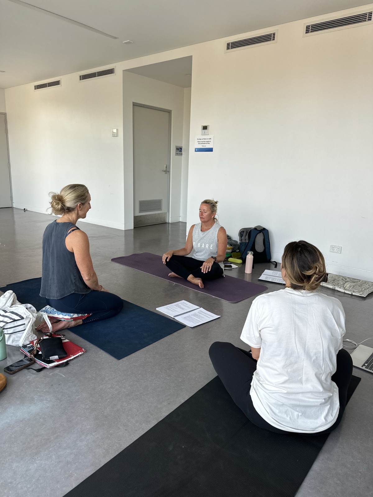 Trainee yoga teachers practising teaching in a small group, with course manuals open on the floor